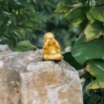 A small golden Buddha statue rests peacefully on a large rock amidst lush green foliage, symbolizing serenity.